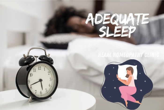 Alarm clock on a bedside table with a person sleeping peacefully in the background, highlighting the importance of adequate sleep for overall health. HEAAL Homeopathy Clinic in Chanda Nagar, Hyderabad offers natural remedies to improve sleep quality.